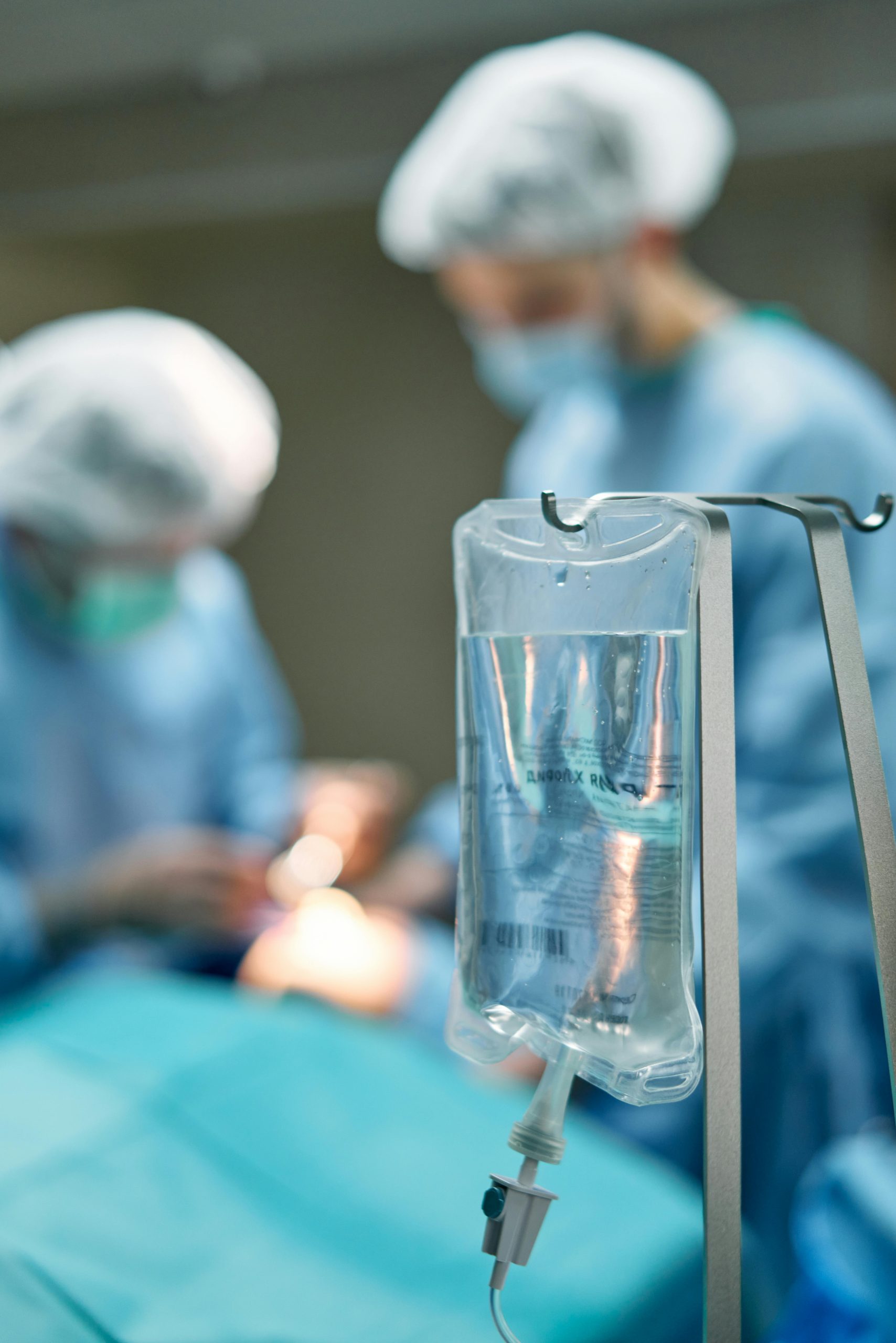 Close-up of an IV drip in a surgical room with medical professionals in the background.